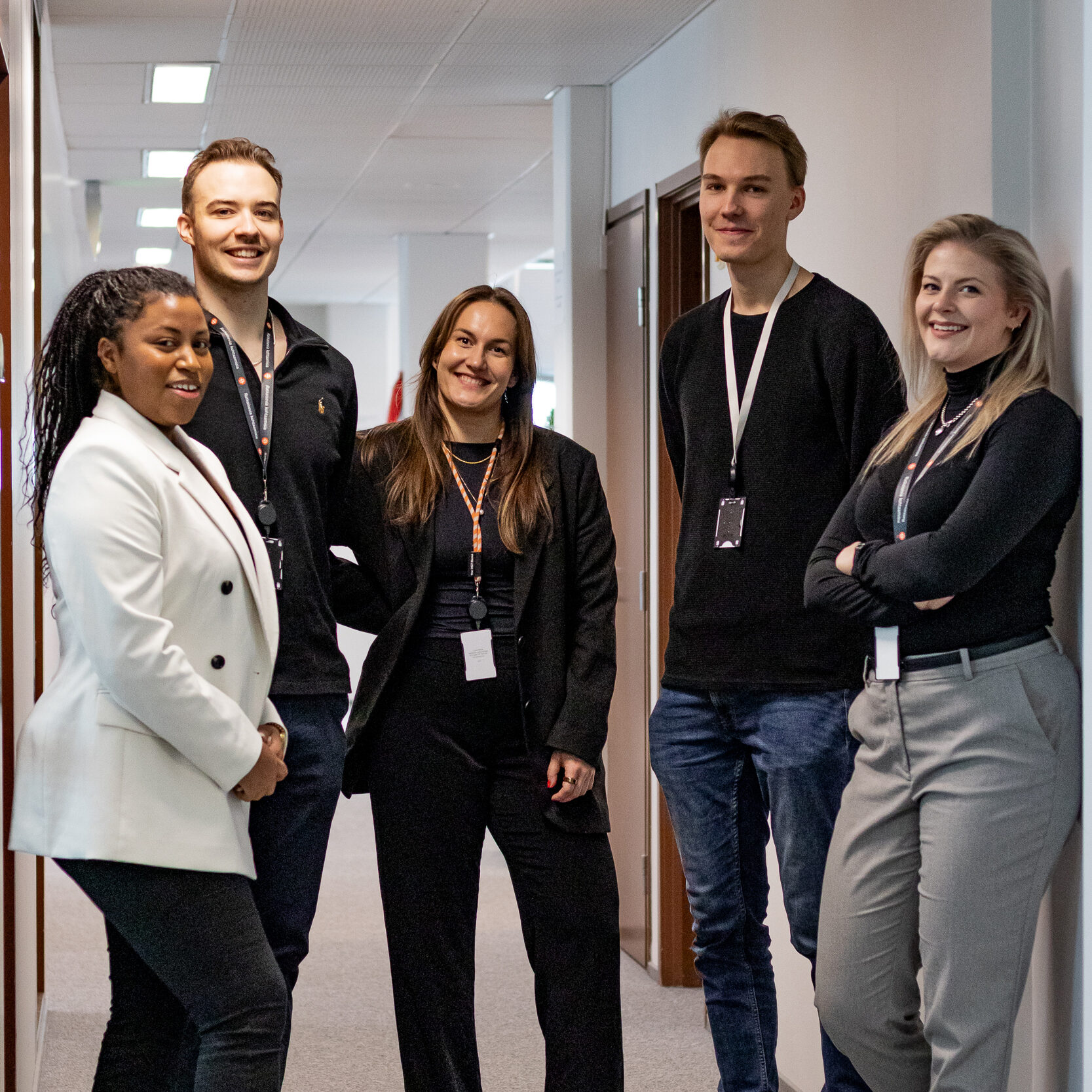 Viisi ihmistä seisoo toimistotilan käytävällä ja katsoo hymyillen kameraan. Tribedo kokemuksia, tiimityötä toimistolla.