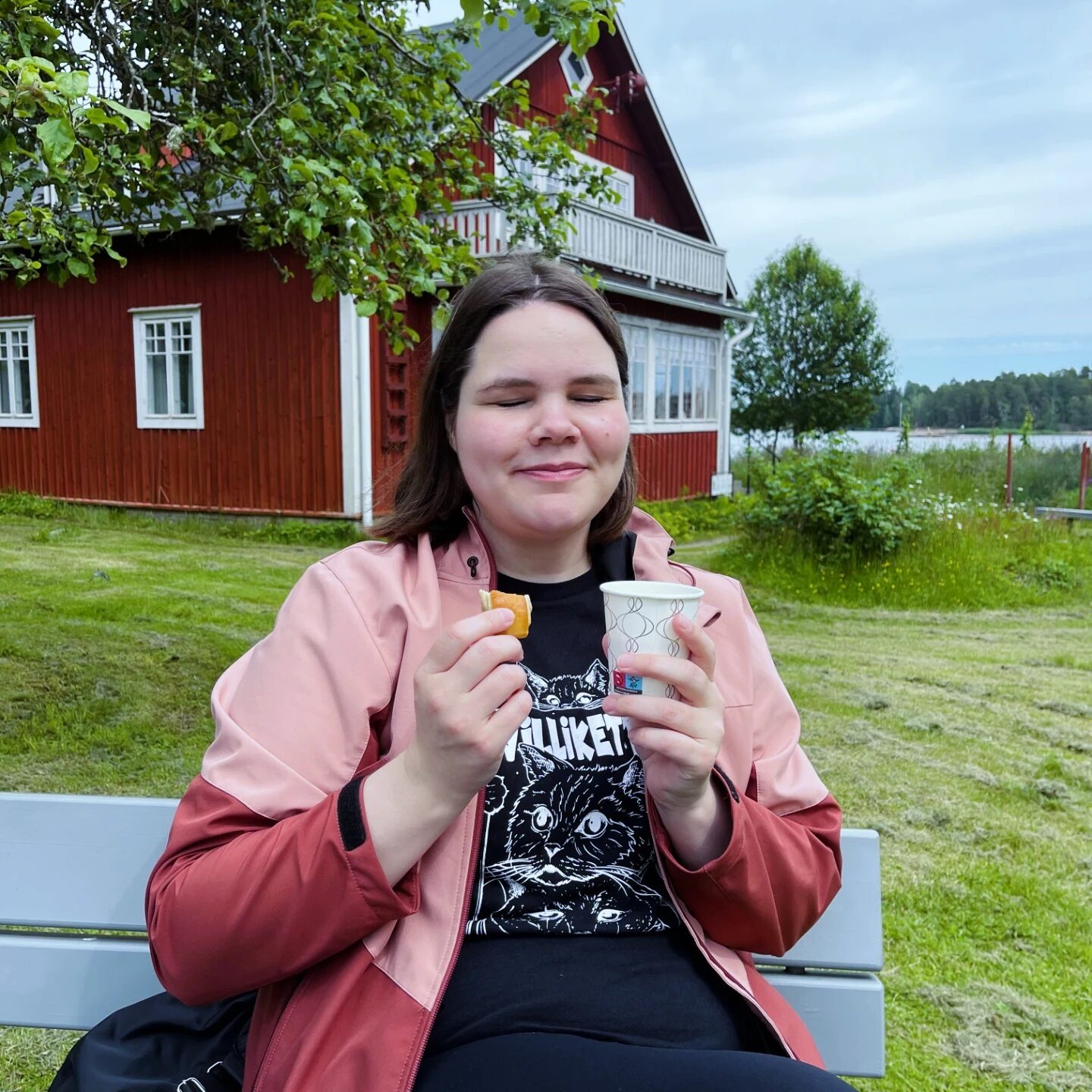 Vuoden tribedolainen 2025 Maria. Istuu ulkona tuolilla, pitäen silmiä kiinni kahvikuppi kädessä. Taustalla punainen talo ja kesämaisema.