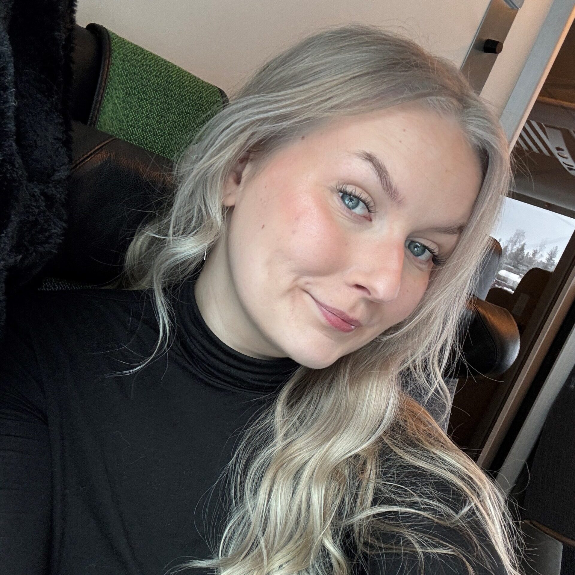 Viestintäagentti 2026 Susanna poseeraa kameralle rennosti hymyillen. Kuvan tausta on junan hytin seinä, ja keskiössä on hänen ilmeikäs katseensa.